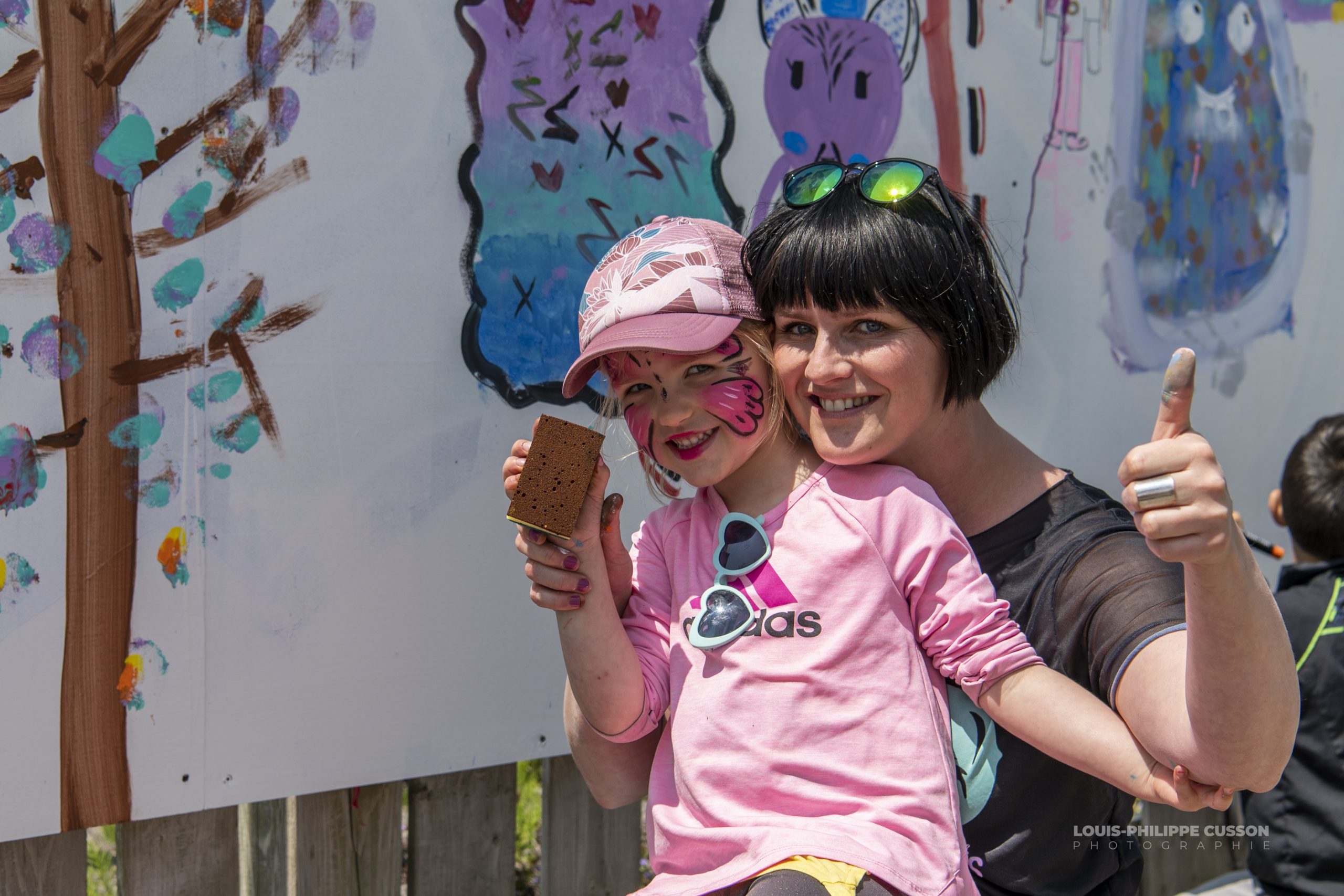 Une enfant au maquillage de papillon et sa mère le pouce levé devant une fresque collective.