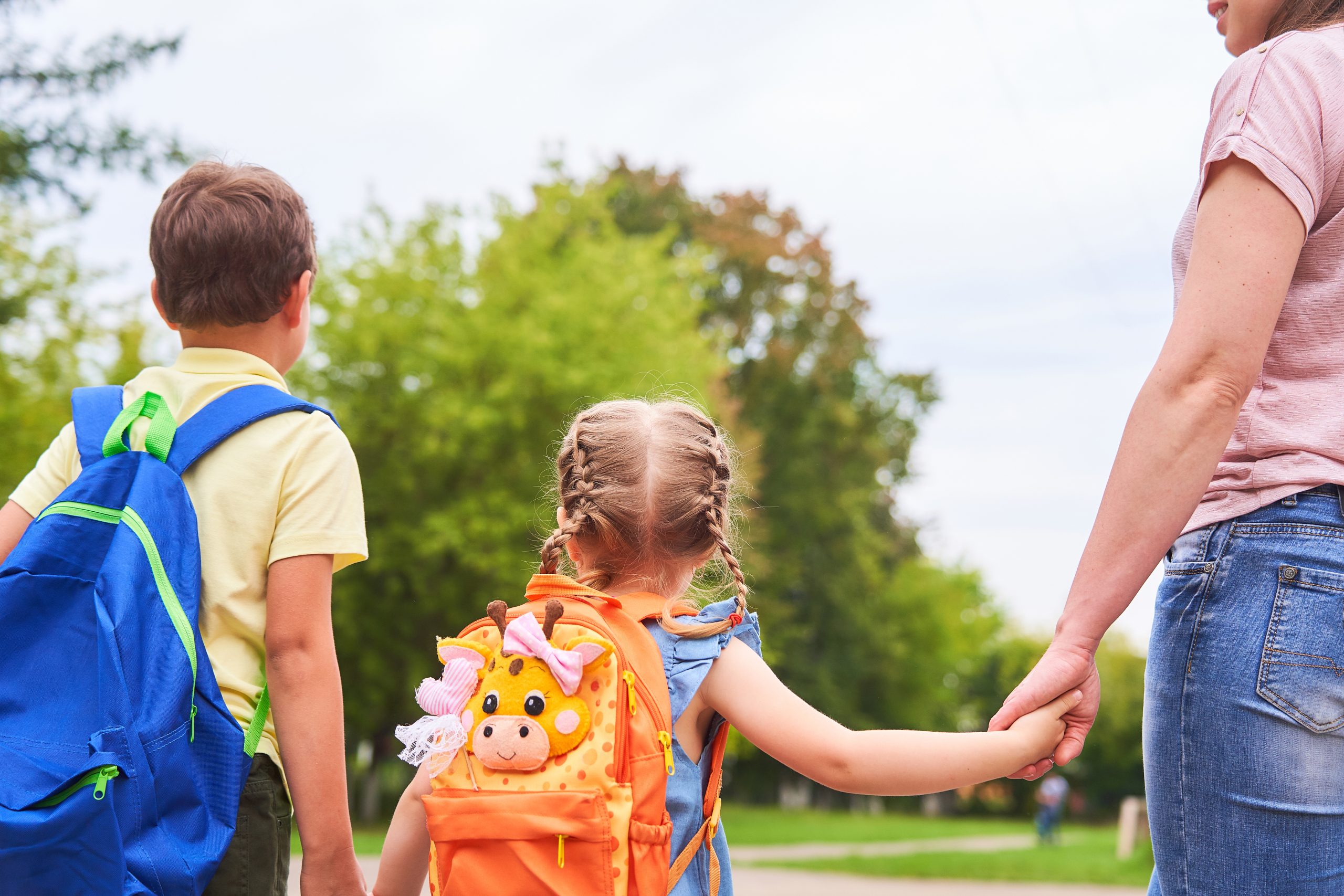 Deux enfants de dos portant leur sac à dos scolaire et leur mère les tenant par la main.